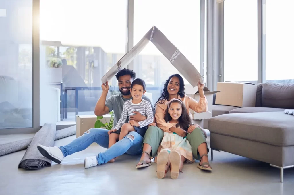 Familia feliz sentada en el suelo de su nueva casa, sosteniendo una caja en forma de techo, rodeada de cajas de mudanza y decoraciones interiores.