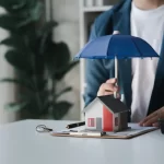 Hombre de negocios sosteniendo un paraguas sobre una casa en miniatura, simbolizando la protección de inversiones inmobiliarias, con documentos y llaves en una mesa blanca.