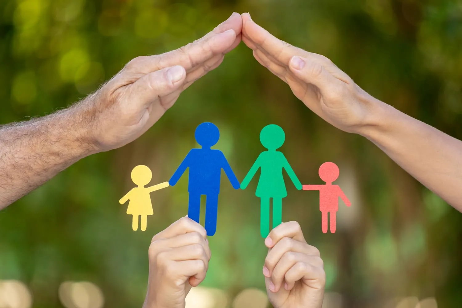 Manos protegiendo figuras de papel que representan a una familia, simbolizando amor y unidad familiar en un entorno natural.